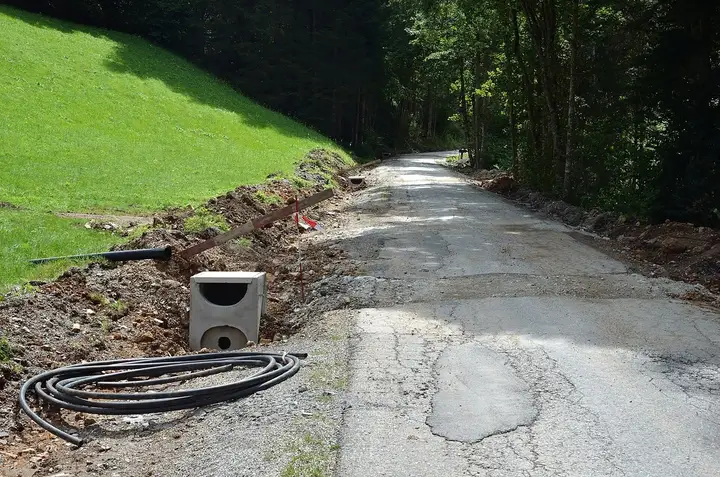 Regard d'avaloir en béton posé au bord d'une route de montagne en travaux, avec une gaine noire enroulée au sol et un tuyau annelé