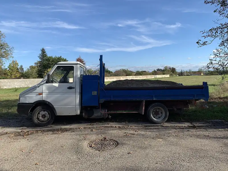 Petit camion benne bleu et blanc chargé de terre, garé en bord de route devant un champ