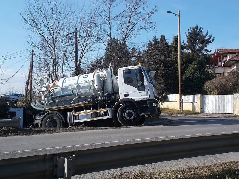 Camion hydrocureur blanc à citerne garé en bord de route, cabine vide, tuyaux d'aspiration souples à l'arrière