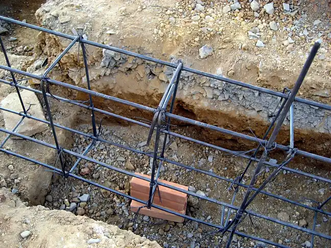 Cage d'armatures en acier posée dans une tranchée de semelle creusée dans un sol caillouteux, cale en brique sous les barres