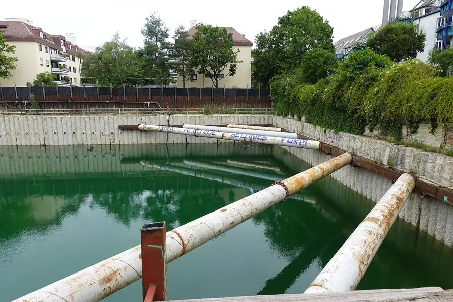 Grande fouille blindée remplie d'eau verte, avec buses tubulaires qui la traversent, immeubles en arrière-plan