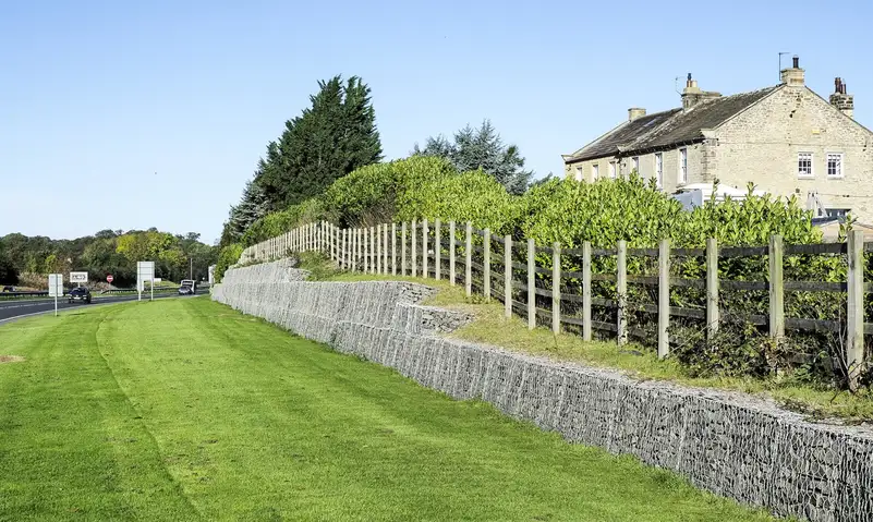 Mur de gabions en gradins retenant un talus planté d'une haie, le long d'une route avec pelouse au premier plan