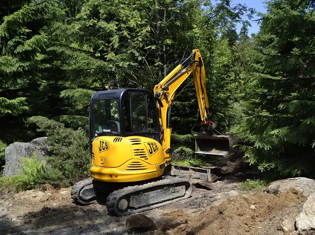 Mini-pelle jaune sur chenilles dans une clairière de sapins, godet relevé, cabine vide, terre remuée au premier plan