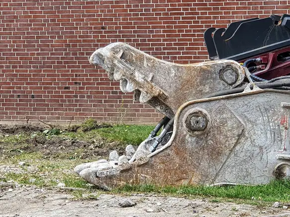 Mâchoires ouvertes d'une pince de démolition posée dans l'herbe devant un mur de briques