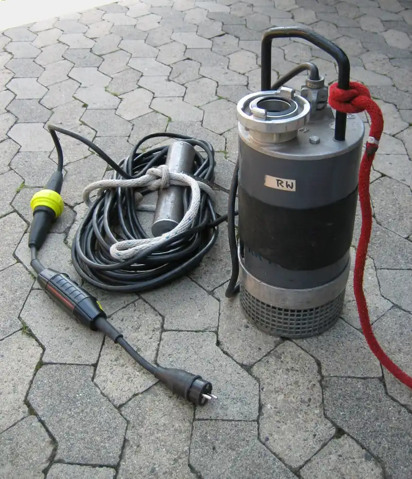 Pompe immergée de chantier grise avec câble enroulé, posée sur un sol pavé