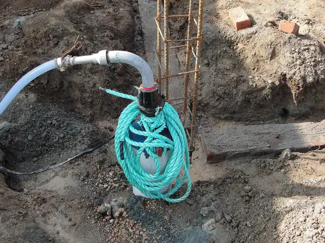 Pompe immergée enroulée d'une corde verte dans une fouille de fondation, tuyau de refoulement blanc, ferraillage à côté