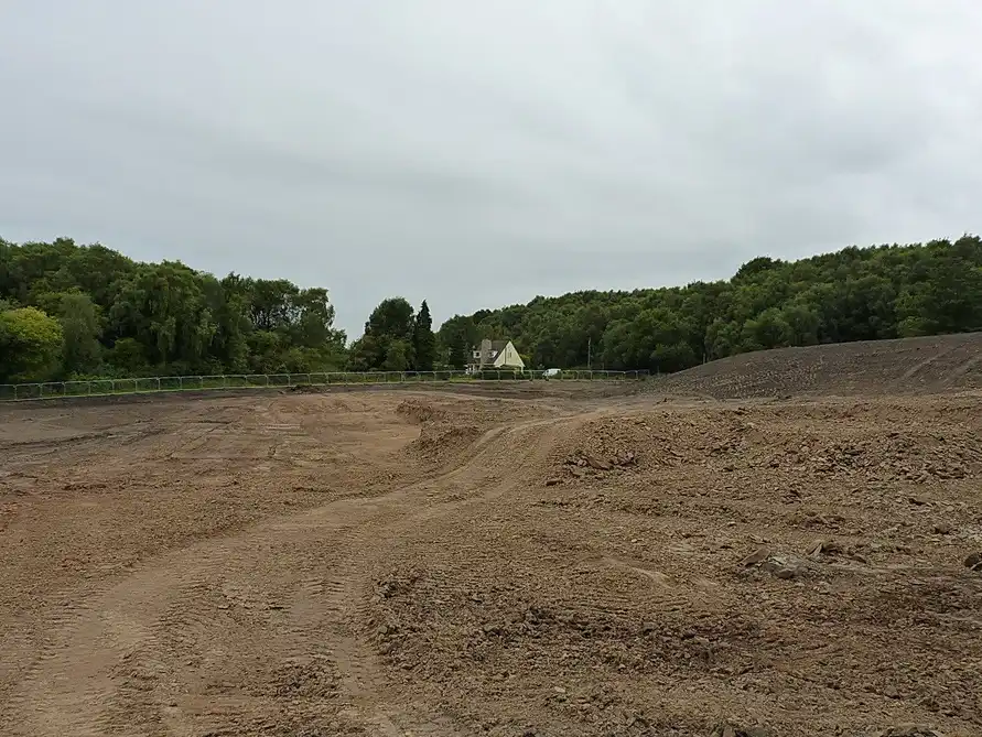 Grand terrain décapé et aplani, terre brune et traces d'engins, haie de clôture et arbres au fond sous un ciel couvert